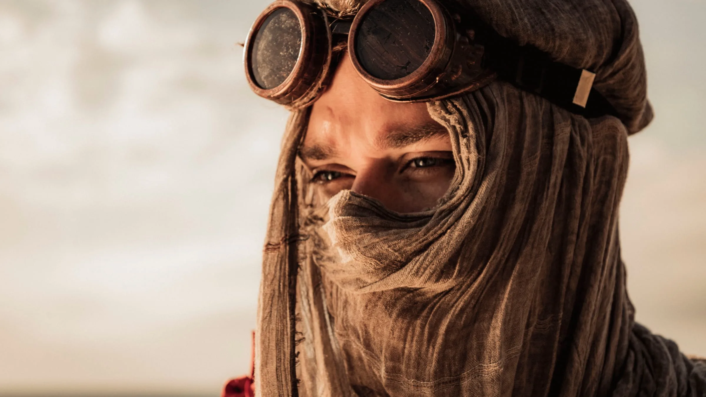 Close-up of a young man with his face wrapped in sand-coloured gauze. Only his eyes show beneath the copper goggles as he looks out at a pale, empty sky to the left.