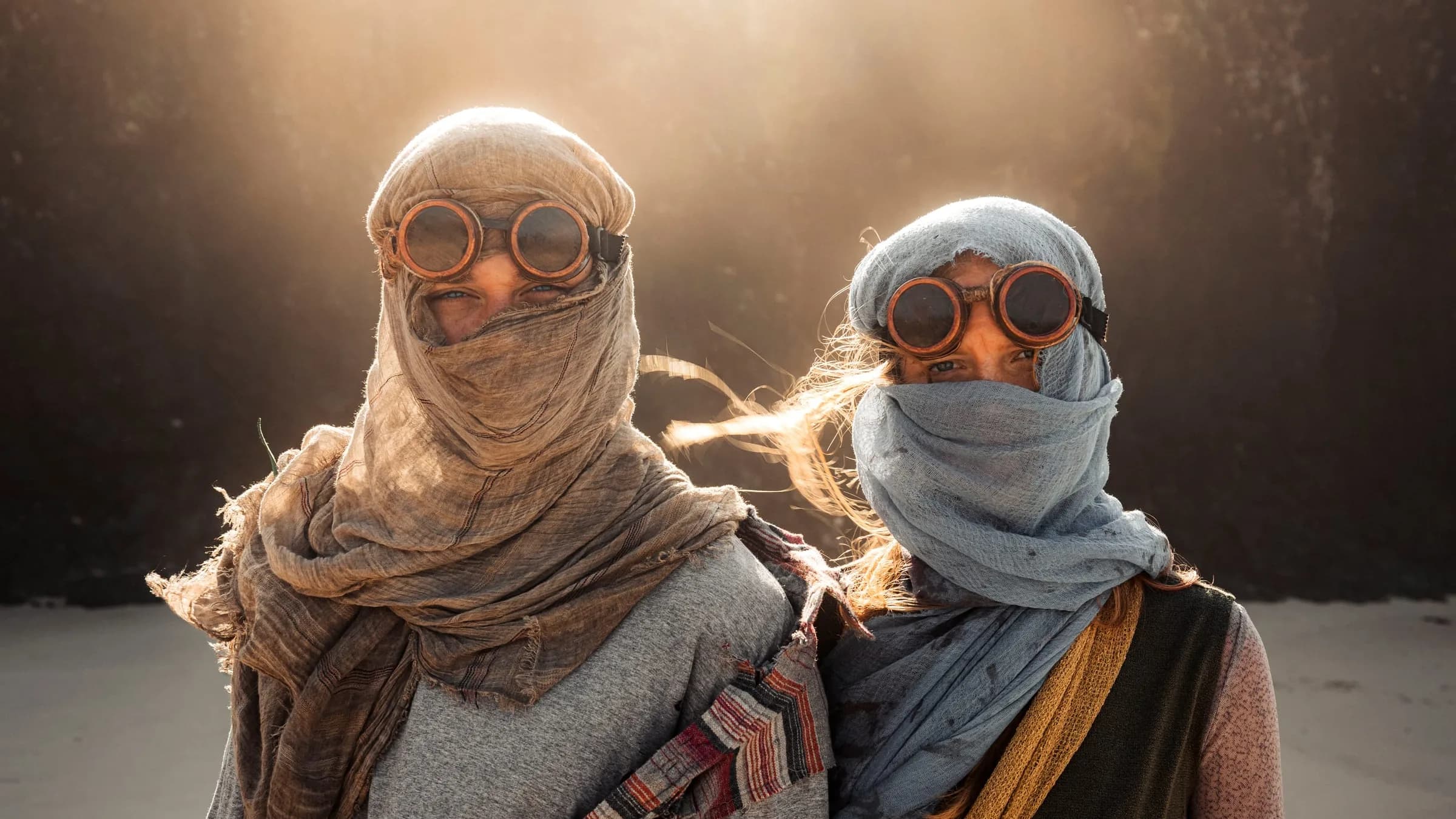 Two young people stand shoulder to shoulder on the sand and look into the camera, faces covered with gauze and copper goggles. Behind them warm haze glows against a dark wall.