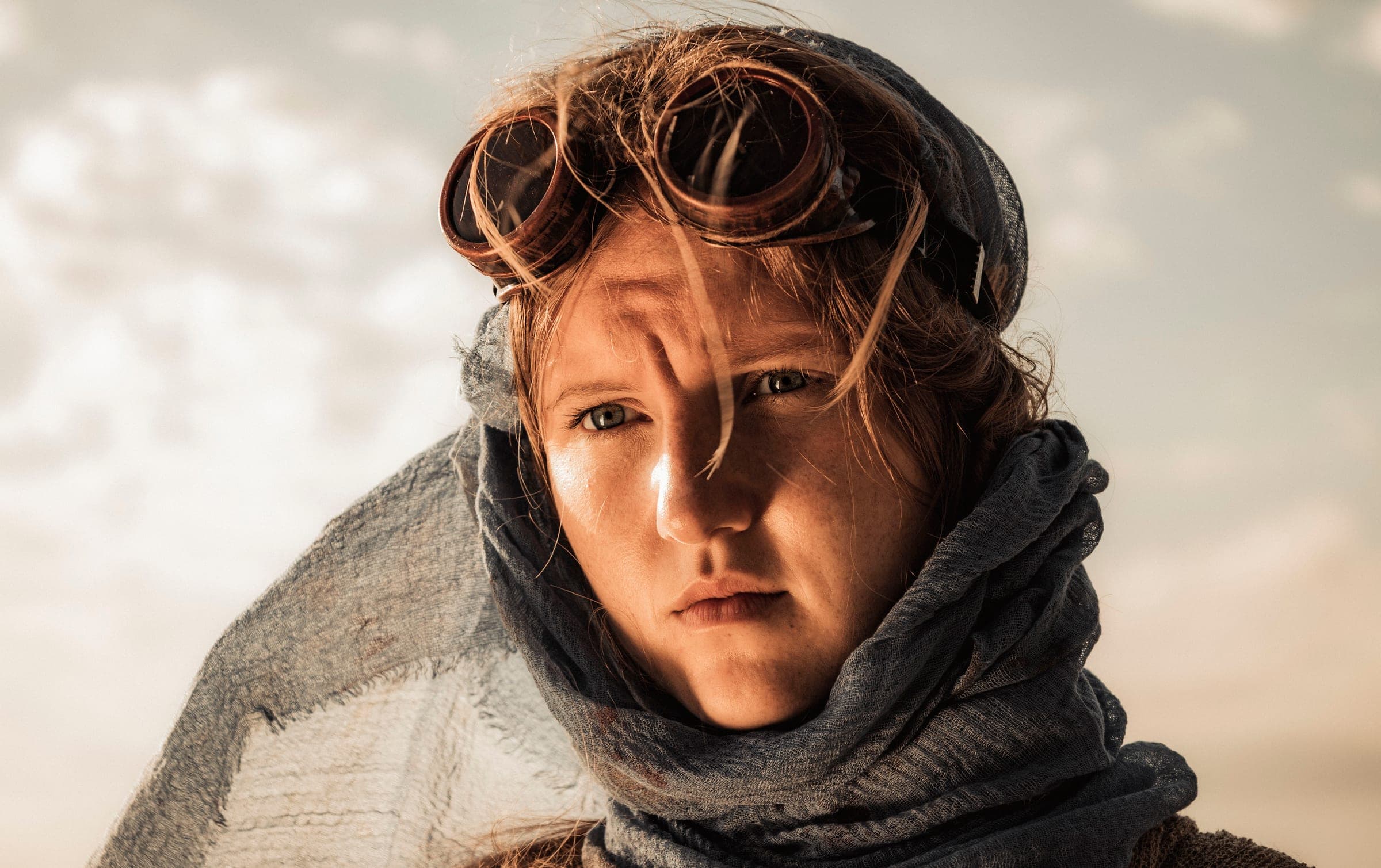 A young woman with auburn hair and freckles looks straight into the camera. Copper goggles are pushed up on her head, a blue-grey scarf is wrapped around her neck, and hard side light casts a shadow across her face.
