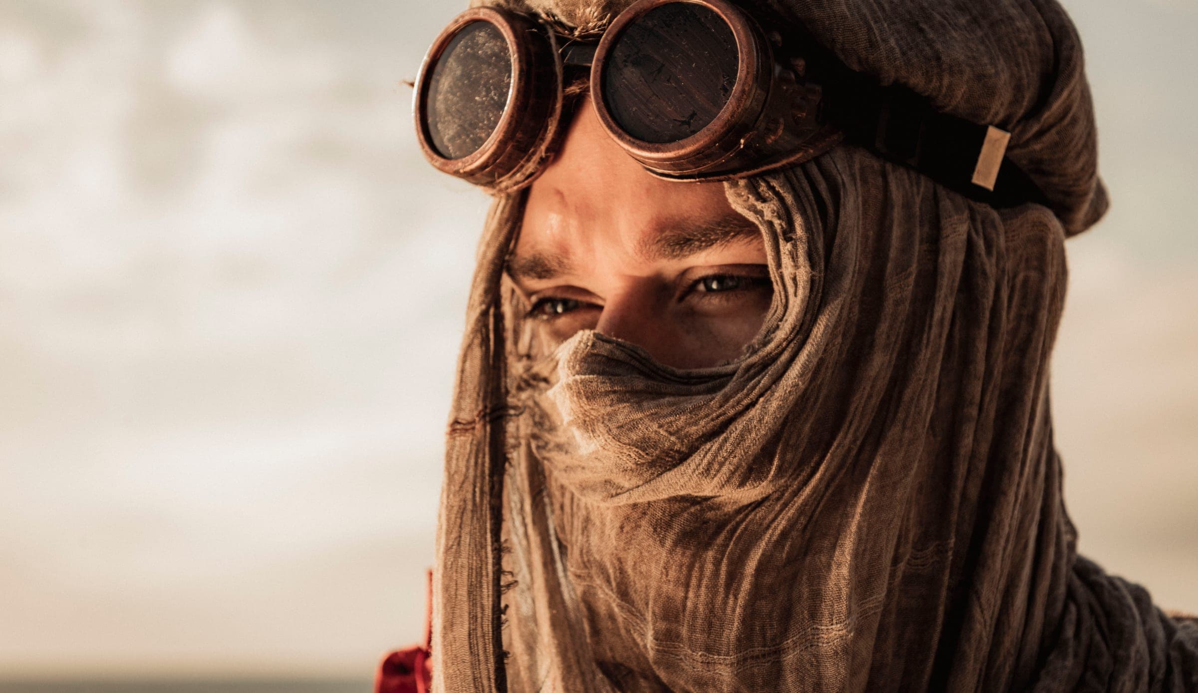 Close-up of a young man with his face wrapped in sand-coloured gauze. Only his eyes show beneath the copper goggles as he looks out at a pale, empty sky to the left.