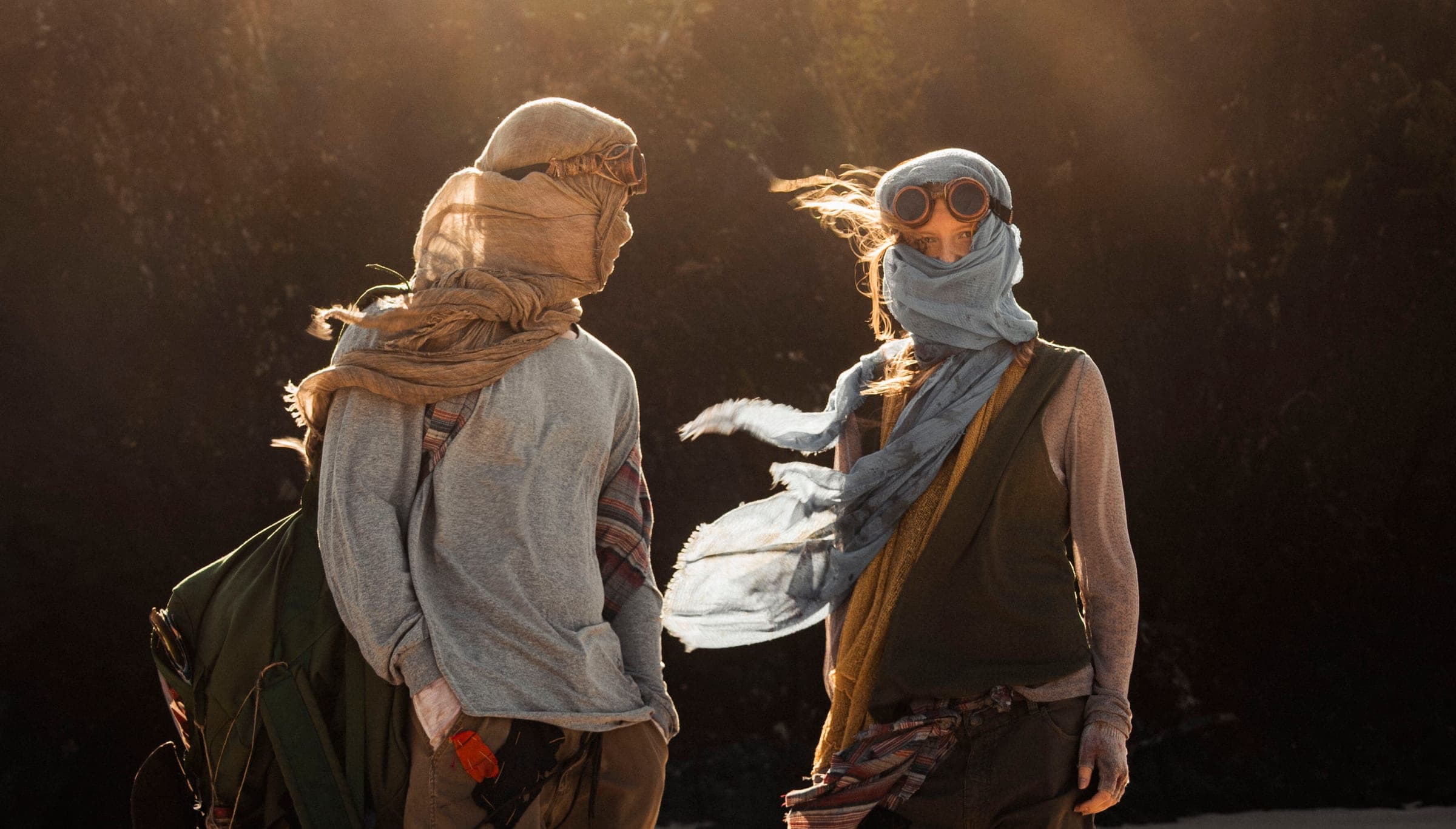 Two young people face each other in front of a dark wall as a gust of wind whips the blue scarf of the woman on the right.