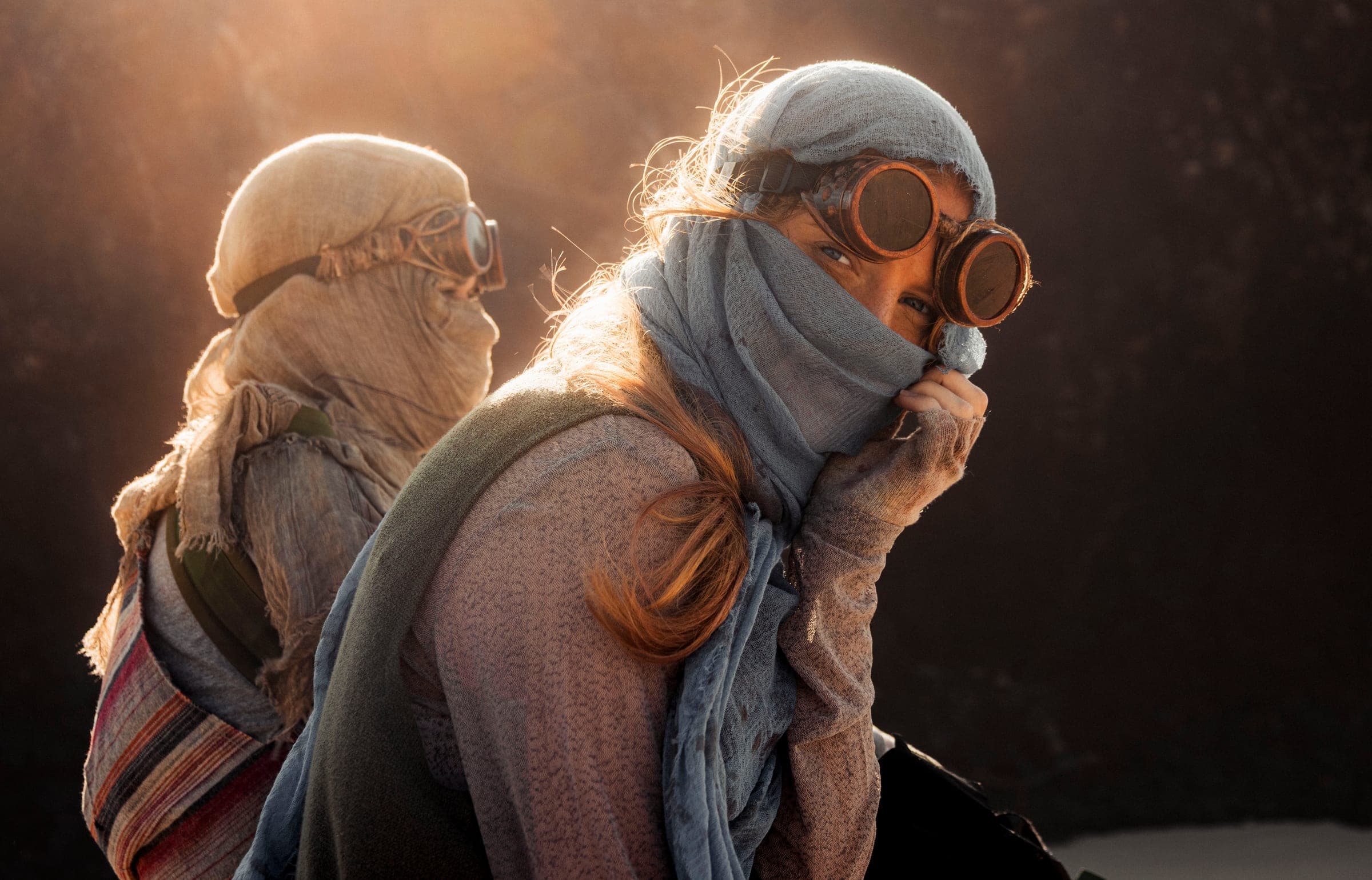 A young woman looks back over her shoulder at the camera, holding a blue scarf over her face. Orange backlight sets her hair aglow, and a blurred figure in gauze stands behind her.