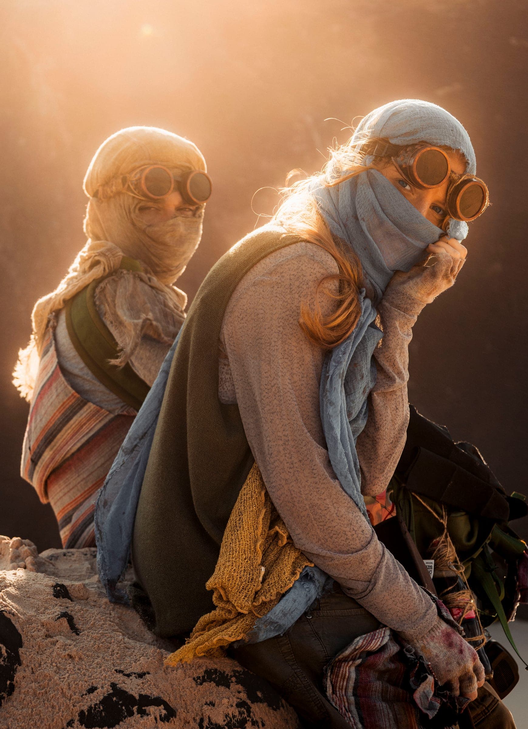 Two young people sit on a rock in golden backlight. The woman in front looks into the camera over her scarf, wearing copper goggles and a mustard knitted scarf. Behind her sits a figure wrapped in gauze.